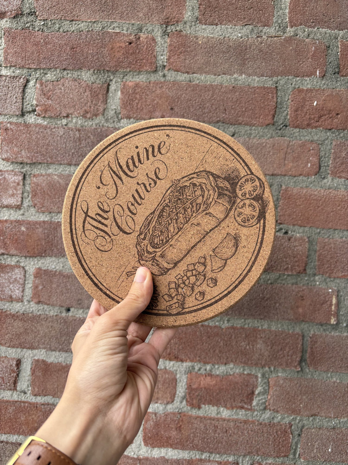 Kurken pannenonderzetter gegraveerd met een gerecht en de tekst The Maine Course, in de hand tegen een bakstenen muur