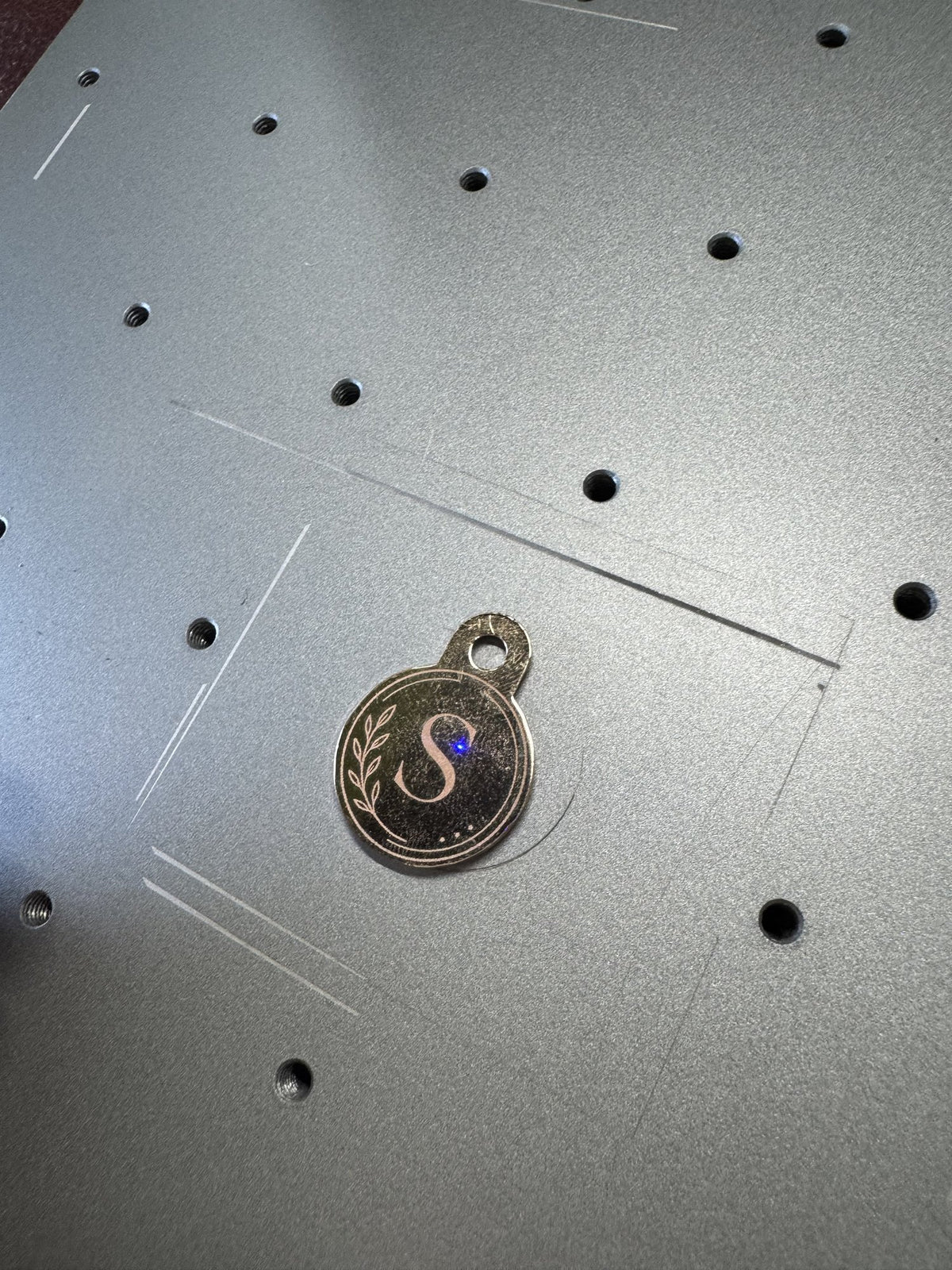 Ronde huisdierpenning van roestvrij staal met een gegraveerde letter S in een lauwerkrans, op het graveerbed van de lasergraveermachine