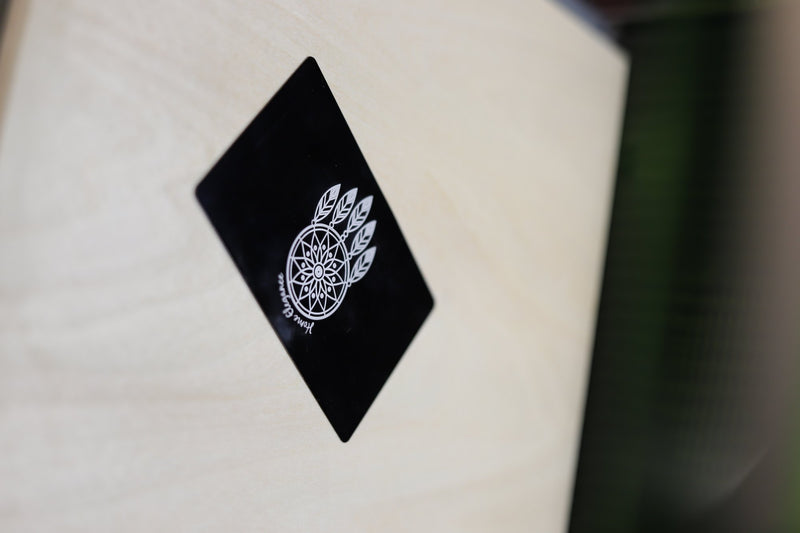 Zwart metalen visitekaartje met een gegraveerd dromenvanger-logo en de tekst Home Elegance, schuin op een houten ondergrond