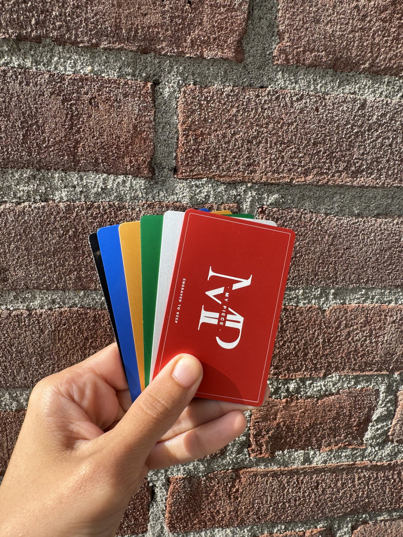 Waaier van zes metalen visitekaartjes in zwart, blauw, goud, groen, zilver en rood, met een gegraveerd logo, in de hand tegen een bakstenen muur